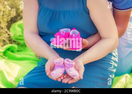 Schwangere Frau und ihr Mann sitzen im Park und halten rosa Babystiefel und Socken in den Händen. Nahaufnahme. Familie, Schwangerschaft, Liebeskonzept, BA Stockfoto