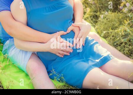 Schwangere Frau und ihr Mann sitzen im Park und halten ihre Hände auf ihrem Baby Beule. Hände in Herzform. Nahaufnahme. Familie, Schwangerschaft, Liebe Stockfoto