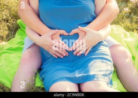 Schwangere Frau und ihr Mann sitzen im Park und halten ihre Hände in Herzform auf ihrem Baby Beule. Nahaufnahme. Familie, Schwangerschaft, Liebe concep Stockfoto