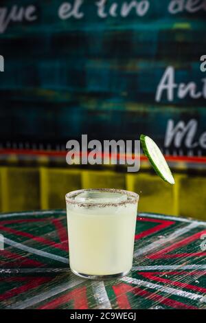 Eine Scheibe Gurke, die in einen Cocktail in einem rocks lowball Glas mit einem großen Eiswürfel fällt, würziger Rand auf dem Glas Stockfoto