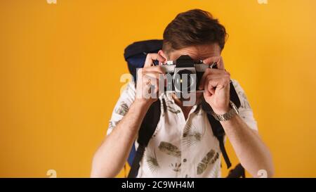 Kaukasischer Reisender mit Rucksack Blick durch den Sucher auf seiner Vintage-Kamera. Isoliert auf orangefarbenem Hintergrund. Hochwertige Fotos Stockfoto