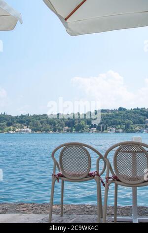 Weiße Bistromöbel am Ufer des Gardasees in Salo, Italien. Die Aussicht lädt zum Entspannen im Schatten ein. Trinken Sie einen Espresso oder ein kühles Getränk. Stockfoto