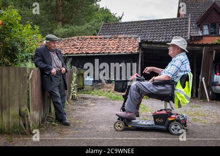 Zwei ältere Männer chatten, einer ist auf einem Mobilitätsroller. Suffolk Village, Großbritannien. Stockfoto