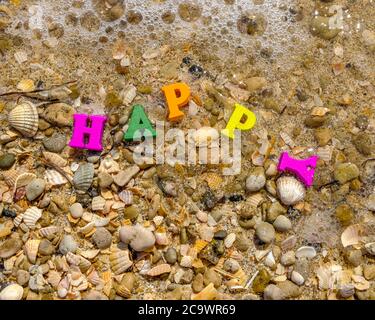 Draufsicht auf Sand und Muscheln, die vom Meerwasser gewaschen wurden. Auf dem Sand, bunte Buchstaben bilden die Inschrift glücklich. Das Konzept von Glück und Stockfoto