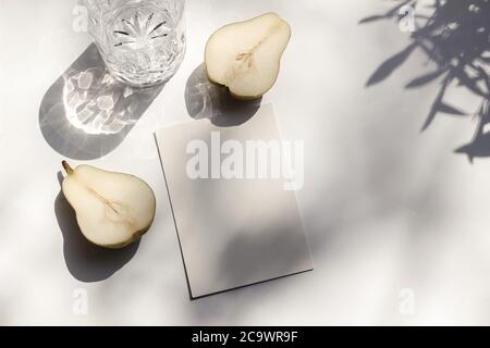 Sommer Schreibwaren Stillleben Szene. Glas Wasser und schneiden Birnenfrucht in Sonnenlicht. Weißer Tisch. Leere Papierkarte, Einladung Mockup Szene, oliv Stockfoto