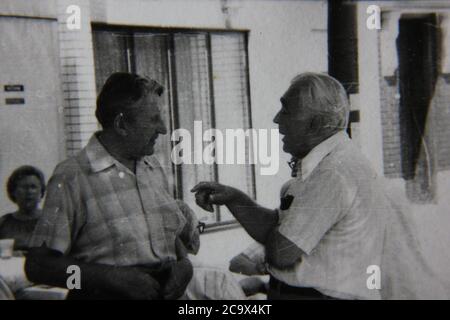 Feine Schwarz-Weiß-Vintage-Fotografie aus den 1970er Jahren von zwei älteren Männern, die eine hitzige Diskussion auf der lokalen Straßenmesse im Freien führen. Stockfoto