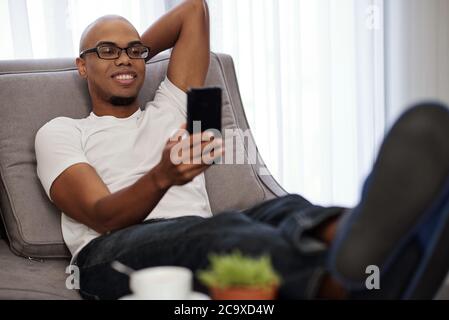 Schöner junger lächelnder schwarzer Mann, der sich zu Hause auf dem bequemen Sofa entspannt und Selfie nimmt Stockfoto