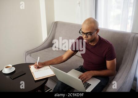 Hübscher junger Mann, der Webinar von Laptop-Bildschirm und Notizen im Notizblock ansieht Stockfoto