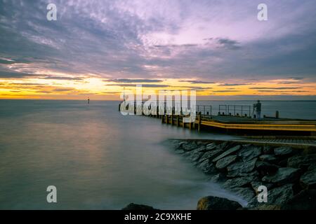 Langzeitbelichtung von seidig glattem Meerwasser und Pier bei Sonnenuntergang mit Fotograf Silhouette und Stativ Stockfoto