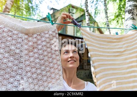 Offen real life Porträt von jungen Erwachsenen schön attraktiv kaukasischen Frau hängen frisch gewaschen Familie Kleidung auf Birke Baum Wäscheleine mit Stockfoto