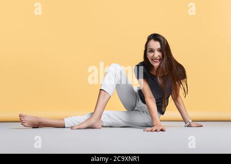 Happy barfuß junge Frau in lässigen Hosen und Hemd sitzt auf dem Boden im Studio auf gelbem Hintergrund Stockfoto