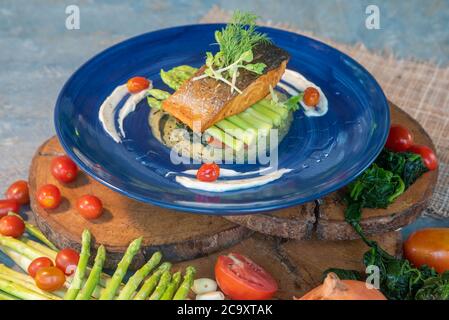 Lachssteak auf Spargel in einem blauen Teller, umgeben von frischen Tomaten und grünen Produkten Stockfoto