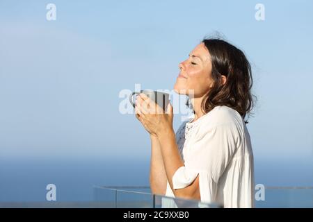 Seitenansicht Porträt einer zufriedenen erwachsenen Frau riechende Kaffeetasse auf einem Balkon am Strand stehen Stockfoto