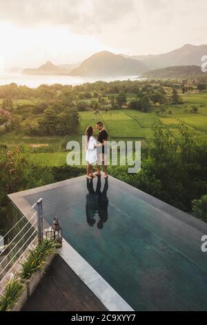 Verliebtes Paar, das am randlosen Pool steht, mit einer tollen Aussicht auf Reisterrassen, Berge und die Küste des Ozeans bei Sonnenuntergang. Luftaufnahme. Flitterwochen in Asien. Stockfoto