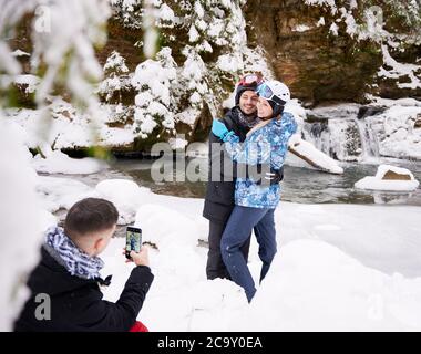 Momentaufnahme des romantischen Paares auf Handy von einem männlichen Freund gefangen. Schöne junge heterosexuelle Paar umarmen, posiert auf malerischen Winter Naturlandschaft Stockfoto