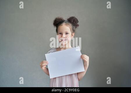 Ein schönes lustiges Mädchen lächelt mit einem weißen Blatt Papier auf einem grauen Hintergrund. Stockfoto