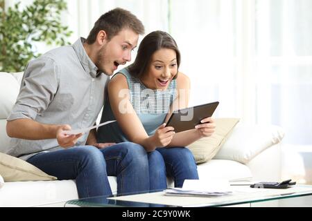 Aufgeregtes Paar, das Tablet und Quittungen überprüft, sitzt auf dem Sofa im Wohnzimmer zu Hause Stockfoto