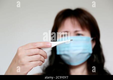 Coronavirus Symptome, Frau in medizinischen Gesichtsmaske misst Körpertemperatur. Besorgt Mädchen schaut auf digitale Thermometer in ihrer Hand Stockfoto