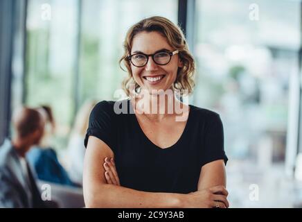 Porträt einer erfolgreichen professionellen Frau mit gekreuzten Armen. Lächelnde Geschäftsfrau, die im Büro steht und die Kamera anschaut. Stockfoto