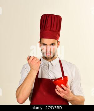 Mann in Kochhut und Schürze hält Werkzeuge. Küchengeräte und Kochkonzept. Koch arbeitet in der Küche. Koch mit besorgtem Gesicht hält Schüssel und Schneebesen auf weißem Hintergrund. Stockfoto