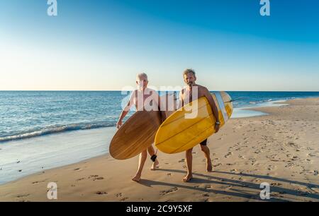 Zwei reife Männer, die mit Surfbrettern am wunderschönen Strand spazieren und das Paradies und den Lebensstil im Ruhestand genießen. Attraktiv fit ältere Erwachsene Freunde Spaß haben Stockfoto