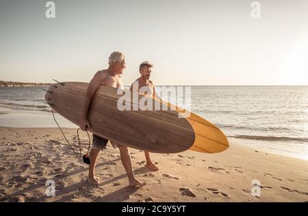 Zwei reife Männer, die mit Surfbrettern am wunderschönen Strand spazieren und das Paradies und den Lebensstil im Ruhestand genießen. Attraktiv fit ältere Erwachsene Freunde Spaß haben Stockfoto