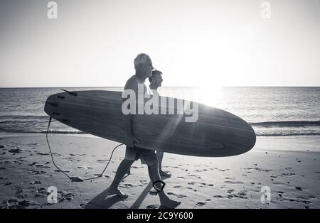 Zwei reife Männer, die mit Surfbrettern am wunderschönen Strand spazieren und das Paradies und den Lebensstil im Ruhestand genießen. Attraktiv fit ältere Erwachsene Freunde Spaß haben Stockfoto