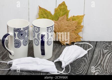 Zwei Glaskeramikbecher. Vor ihnen sind Schutzmasken für die Dauer der Pandemie. Hinter ihnen sind getrocknete Herbst Ahornblätter. Stockfoto