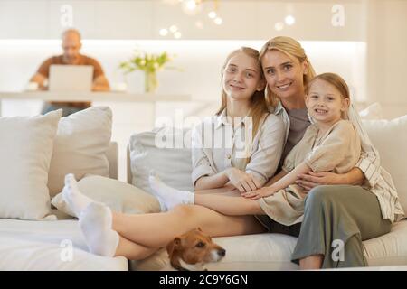 Warm-getönte Porträt von glücklichen Mutter umarmt zwei Töchter, während posiert zusammen auf der Couch sitzen in gemütlichen Haus Interieur mit Vater im Hintergrund, CO Stockfoto