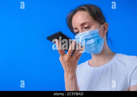 Frau in medizinischen Gesichtsmaske hält Smartphone-Gerät, mit Spracherkennungsfunktion, Aufnahme von Audio-Nachricht, im Gespräch mit mobilen Assistenten Stockfoto