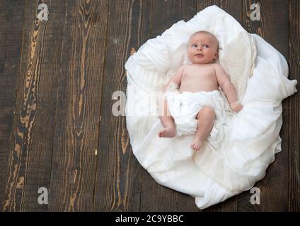 Neugeborenes Kleinkind mit blauen Augen und neugierigen Gesicht auf Holzhintergrund, kopieren Raum. Kleinkind mit weißer Bettdecke bedeckt. Kindheit und Unschuld Konzept. Baby liegt auf weißen weichen Decke als Nest entworfen Stockfoto