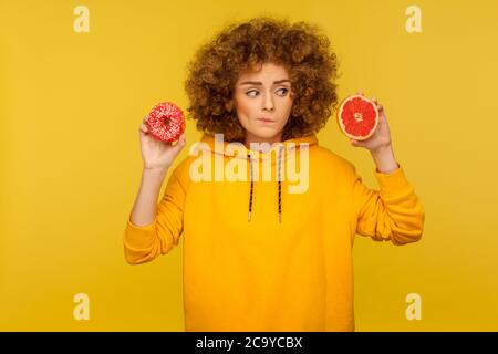 Gesunde Früchte vs kalorienreiche Dessert. Porträt von nachdenklichen, verwirrt lockigen jungen Frau im urbanen Stil Hoodie Wahl zwischen Grapefruit und d Stockfoto
