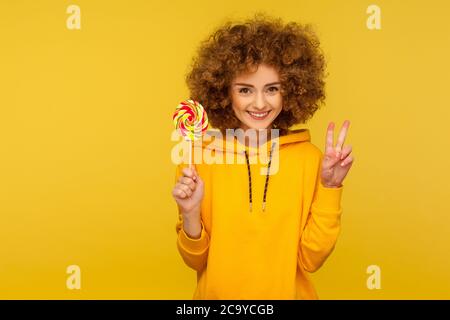 Süße Regenbogenbonbons. Porträt einer glücklichen modernen lockigen Frau im urbanen Stil Hoodie mit Lollipop und Siegesgeste, lächelnd an der Kamera Stockfoto