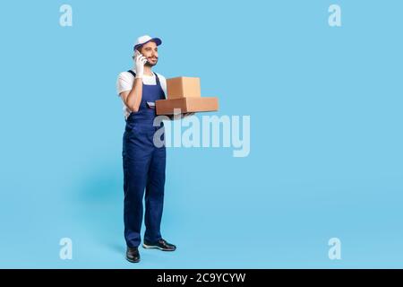 In voller Länge glücklich freundliche Lieferung Mann in Uniform sprechen auf Handy, halten Kartons und Kommunikation mit Kunden. Kurier beantwortet Phon Stockfoto