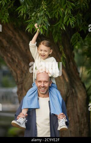 Vertikale Porträt von niedlichen kleinen Mädchen lachen glücklich, während sitzen auf Väter Schultern und genießen Spaziergang im Park, beide Blick auf die Kamera Stockfoto