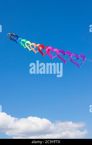 Eine Reihe von regenbogenfarbenen, herzförmigen Drachen, die am blauen Himmel über weißen Wolken fliegen. Hintergrund oder Hintergrundbild. Vertikale Aufnahme. Stockfoto