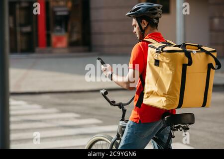 Junge männliche Radfahrer mit Kurier Liefertasche mit Handy auf der Straße, um Adresse des Kunden in der mobilen Anwendung zu suchen Stockfoto