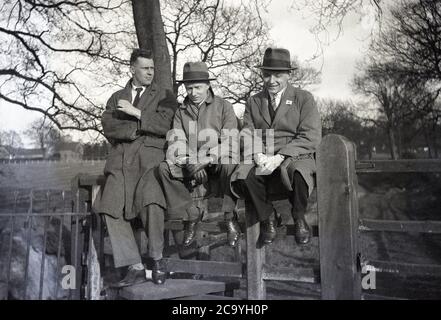 1940er/50er Jahre, historisch, drei elegant gekleidete, stilvolle Männer in Mänteln und zwei mit smarten Trilby-Hüten, zusammen auf einem Holzzaun bei einem Park oder möglicherweise einem Schulspielplatz sitzend, Vielleicht zu einem Land Pferderennen Treffen gewesen und denken, was hätte sein können, wenn ihre Wetten gewonnen hatten, England, Großbritannien. Ein Trilby ist ein schmaler Hut, der manchmal in Großbritannien als 'brauner Trilby' bezeichnet wird und häufig bei Pferderennen gesehen wird. Der Name stammt aus der Bühnenbearbeitung von George du Maurier's Roman Trilby von 1894. Stockfoto