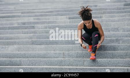 Muskulöse afroamerikanische Frau in Sportbekleidung, mit modernen Kopfhörern, Fitness-Tracker, Schnürsenkel auf Turnschuhen auf Treppen binden Stockfoto