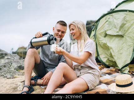 Junge Paar Reisende in lässigen Outfits mit Zelt. Mann gießt Tee aus Thermoskannen. Lokaler Tourismus, Wochenendausflug Stockfoto