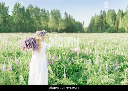 Ein Mädchen mit einem Blumenstrauß über der Schulter in einem weißen Kleid und einem Blumenkranz geht zurück entlang des Blumenfeldes, im Blumenfeld von Stockfoto