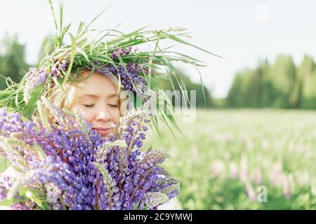 Ein blondes Mädchen in einem Blumenkranz mit einem Blumenstrauß von Lupinen lächelt sanft und inhaliert den Duft von Blumen in einer Blumenwiese auf einem sonnigen Stockfoto