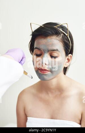 Eine graue Schlammmaske wird auf eine schöne junge Frau aufgetragen. Schönheitsbehandlungen. Gesichtspflege, Poren straffen und feuchtigkeitsspendend mit natürlichen kosmetischen Cremes. Stockfoto
