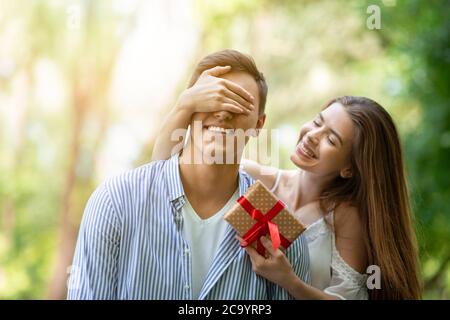 Junges Mädchen schließen die Augen ihres Freundes und überraschen ihn mit Geburtstagsgeschenk im Park Stockfoto