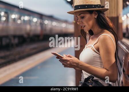 Fröhliche junge hispanische Brünette Frau auf Bank auf Bahnsteig mit Telefon, Hua Lamphong Station, Bangkok, Thailand Stockfoto