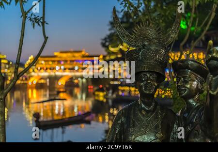 Feng Huang, China - August 2019 : Skulptur der einheimischen ethnischen Minderheit Mann und Frau in der historischen Altstadt beleuchtet in der Nacht, Provinz Hunan Stockfoto