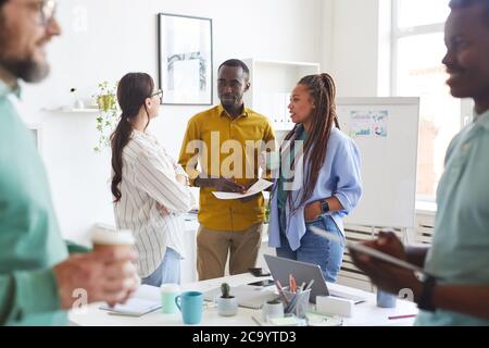 Multiethnische Gruppe von kreativen Geschäftsleuten, die während der Pause im Konferenzraum chatten, konzentrieren sich auf lächelnde afroamerikanische Männer, die mit Frauen in legerer Kleidung sprechen, Kopierraum Stockfoto