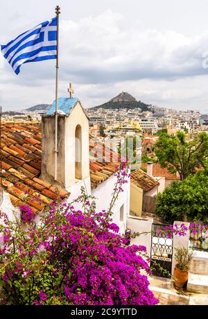 Skyline von Athen, Blick von Anafiotika in Plaka, Griechenland. Plaka ist berühmte Touristenattraktion von Athen. Altes Haus mit Blumen und griechischer Flagge Stockfoto