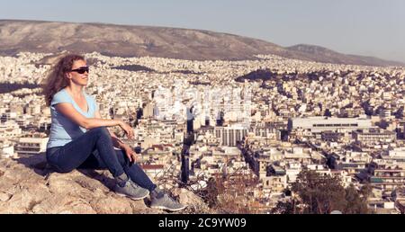 Junge Frau ist auf dem Hintergrund der städtischen Landschaft von Athen, Griechenland, Europa. Erwachsene hübsche Mädchen Tourist entspannt auf einem Hügel mit Blick auf Athen Stadt in Summe Stockfoto