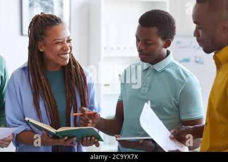 Waist-up-Porträt einer zeitgenössischen afroamerikanischen Frau, die fröhlich lacht, während sie mit Kollegen im Büro über das Projekt spricht Stockfoto